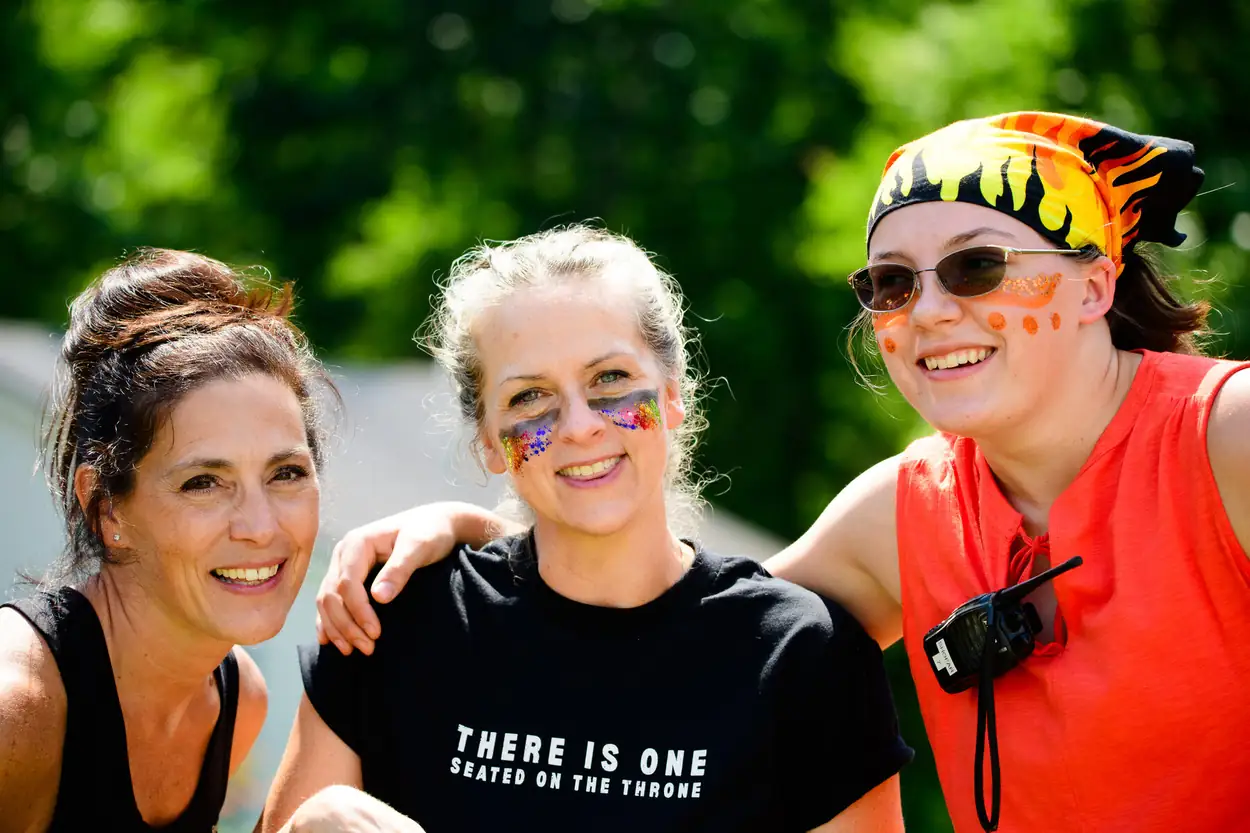 Three women arm in arm with glitter and face paint under their eyes; the woman in the middle wears the camp shirt reading THERE IS ONE, SEATED ON THE THRONE.