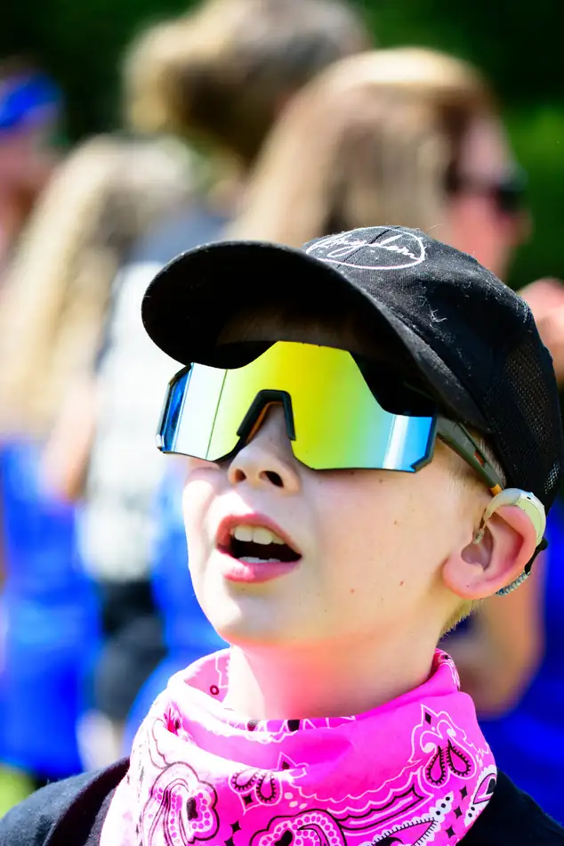A camper in a black cap and rainbow mirrored sunglasses with a hot pink bandana at his neck, mouth open mid-yell.