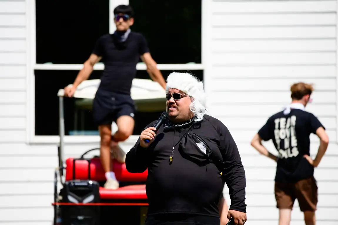 A leader in a long white wig speaking into a microphone outside a white clapboard building, someone standing on a red cart behind him, and at the right a camper with his back to the camera in the camp shirt, TEAR DOWN THE IDOLS stacked in four lines across it.