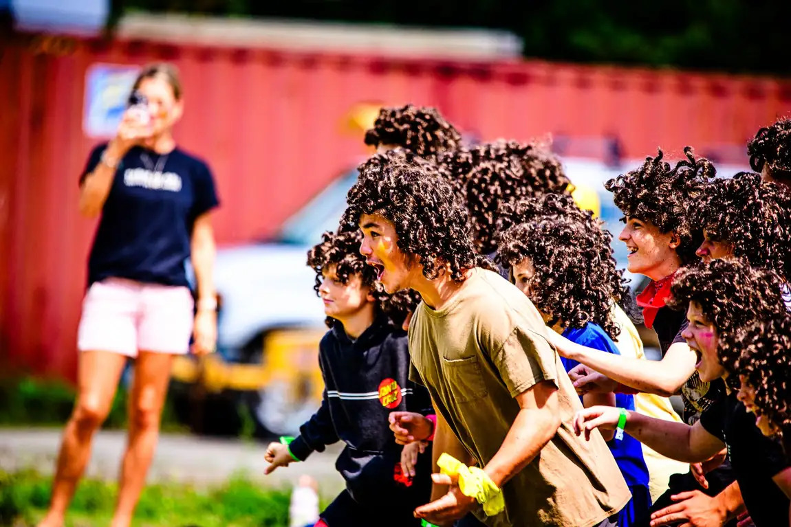 A wall of campers in brown curly wigs charging forward together, mouths open, past a red shipping container.