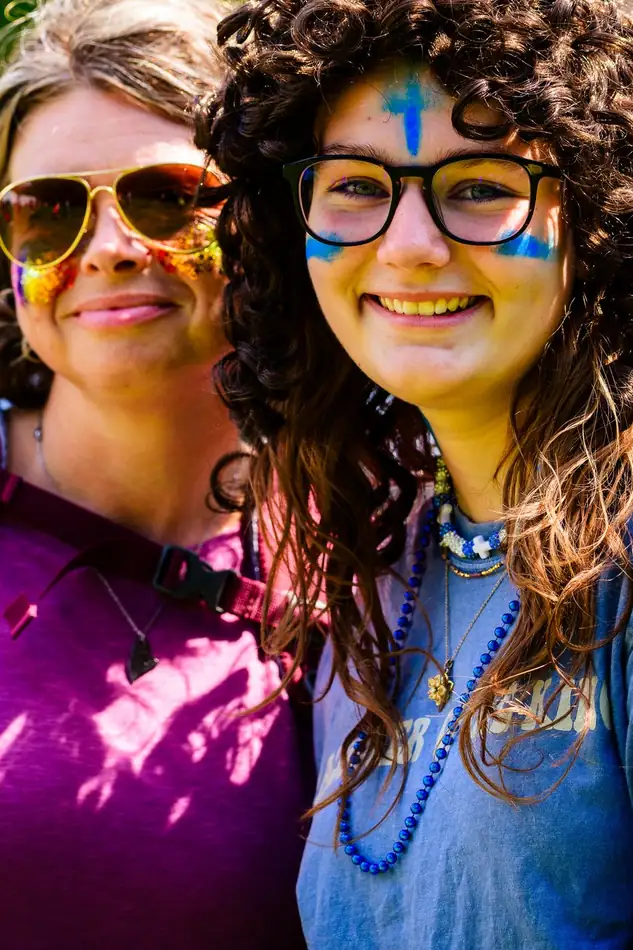 A mother in sunglasses and orange face paint cheek to cheek with her daughter in a curly wig and blue cross paint.
