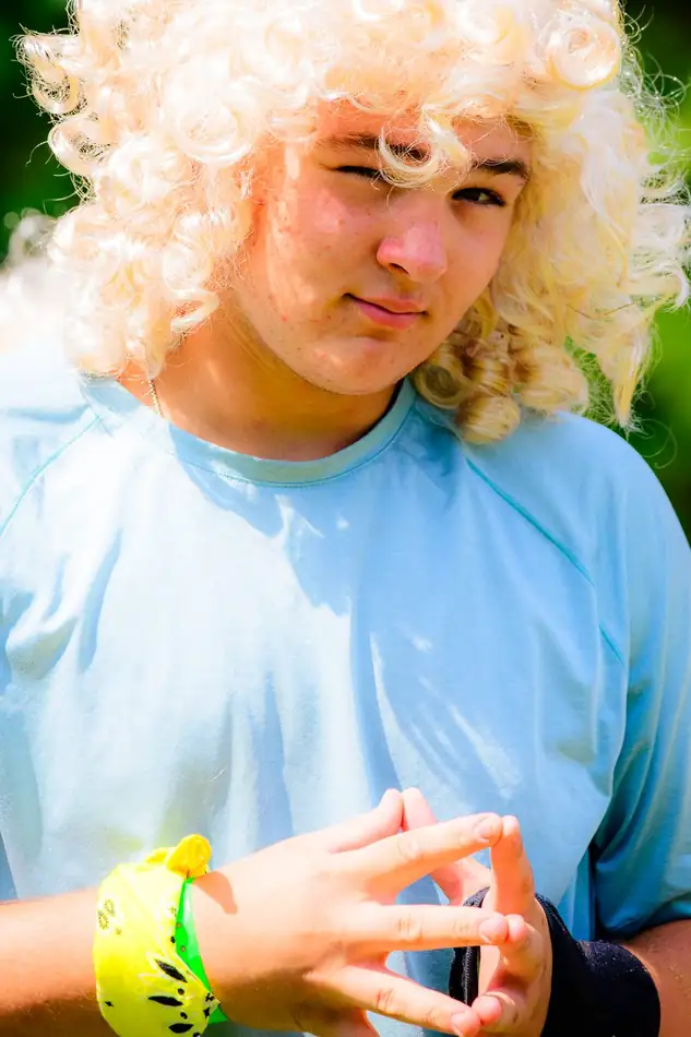 A boy in a blond curly wig and pale blue tee with sun on his face.