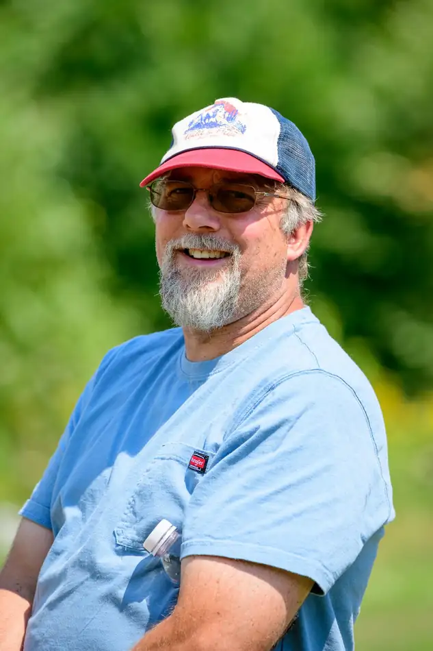 A man with a grey beard, a ball cap and glasses, smiling in a pale blue shirt.