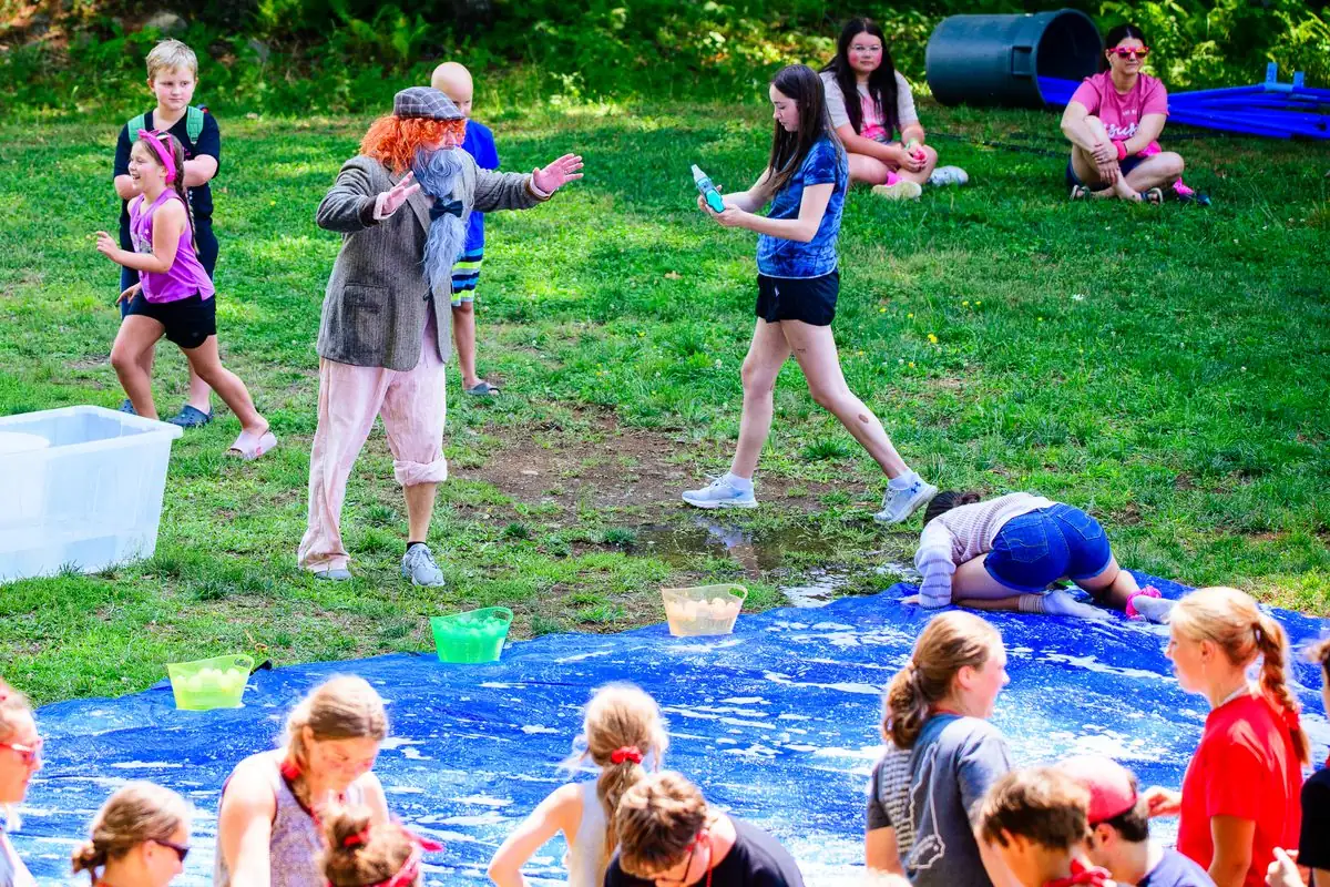 Kids packed along the near edge of a blue tarp on the grass, a leader in an old-man costume with his arms flung wide behind them, a water tub and the pines beyond.