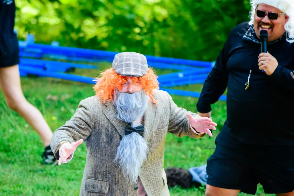 A leader in a full old-man costume with a grey wig and long white beard, arms flung wide, while another leader holds a microphone.
