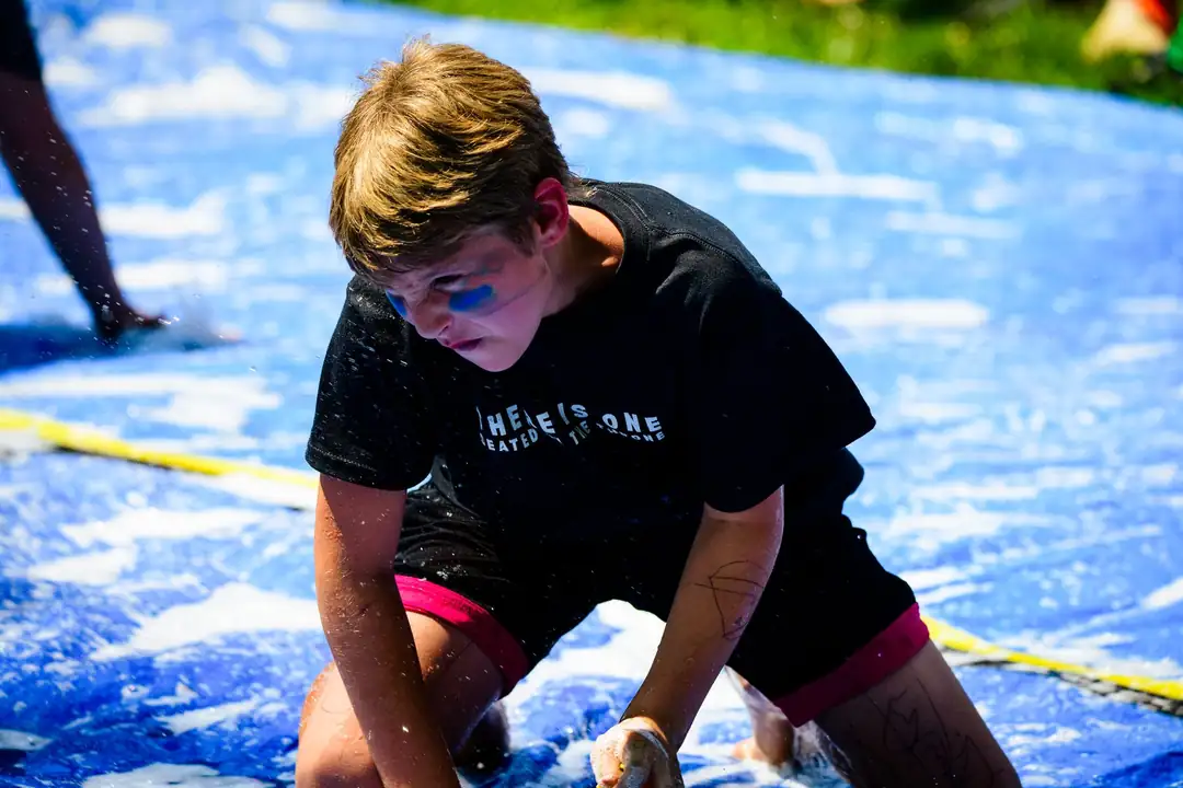 A boy on his hands and knees on a soapy blue slip-n-slide, blue paint on his face.