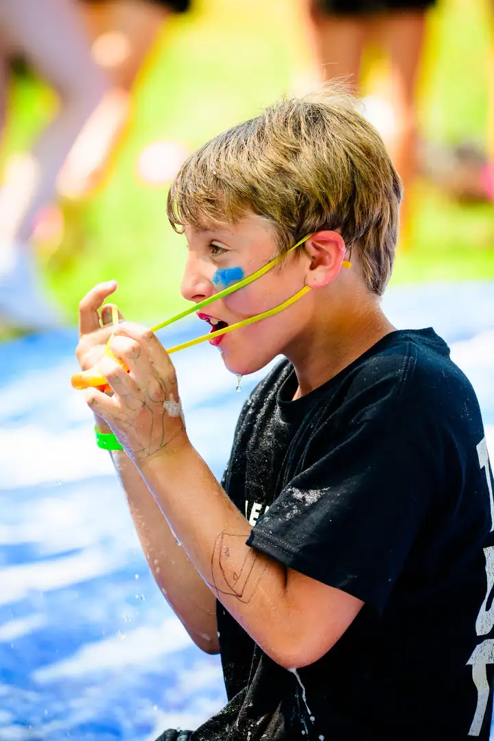 A boy loading a water balloon slingshot with his teeth, cheeks puffed in concentration.