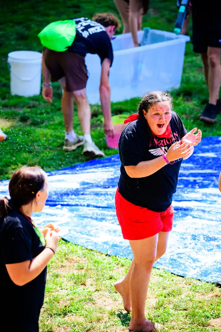 A girl beside the blue tarp with her mouth open mid-shout and her hands up, other campers working around a water tub behind her.