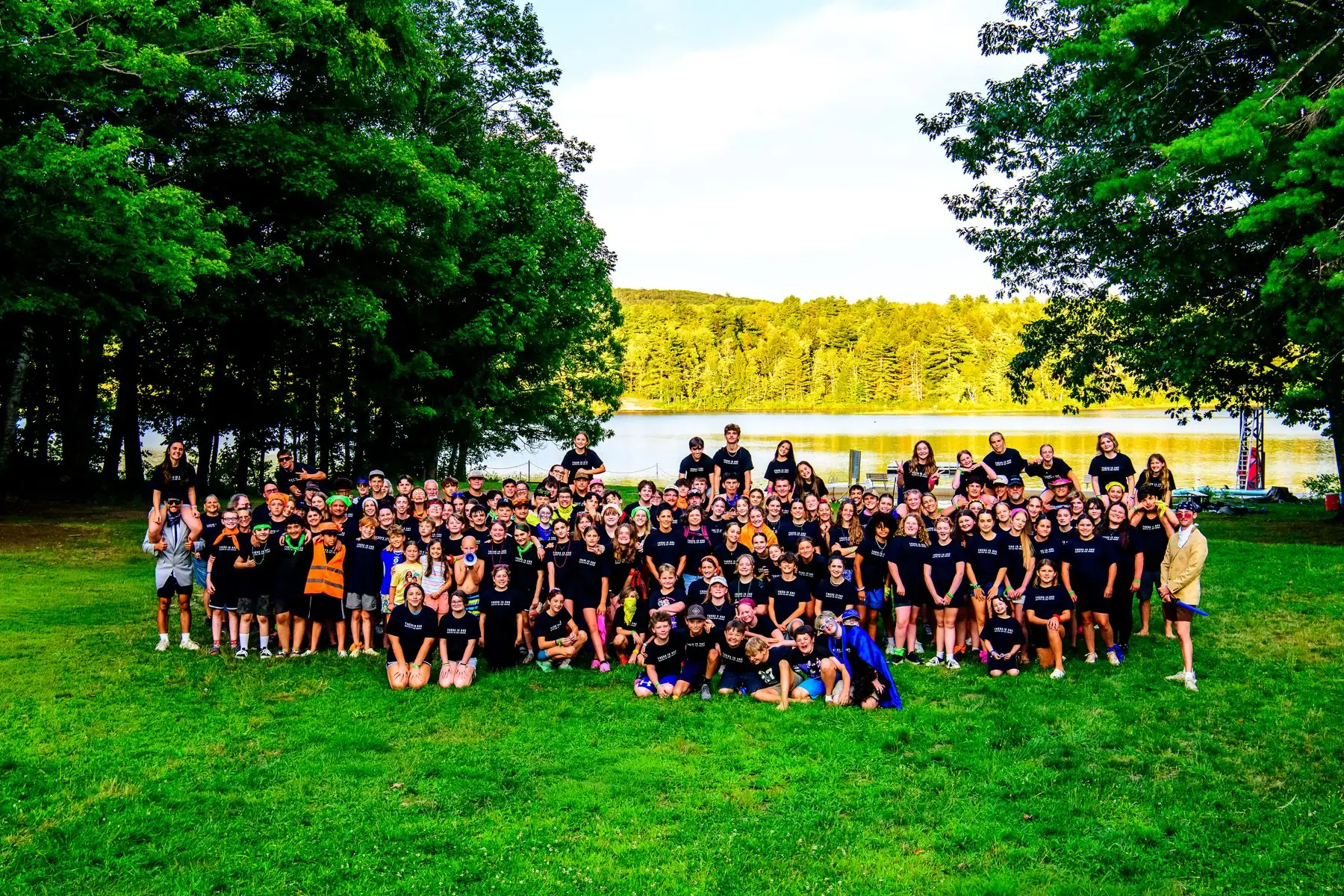 The whole camp gathered on the grass in front of tall pines, the lake and far shore behind them, nearly everyone in a black camp shirt.