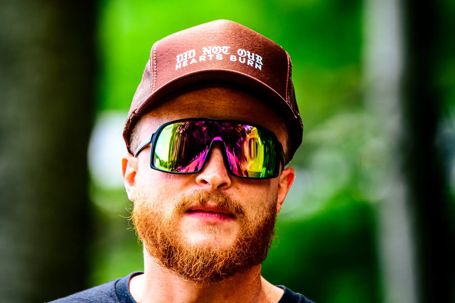 A close crop of a rust-red trucker cap reading DID NOT OUR HEARTS BURN in blackletter and tracked capitals, worn above a red beard and mirrored sunglasses.