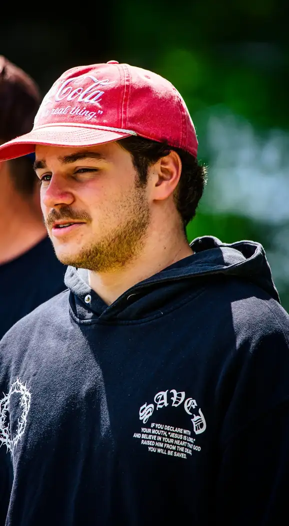 A young man in a faded red cap and a black hoodie with SAVED across the chest in gothic lettering.