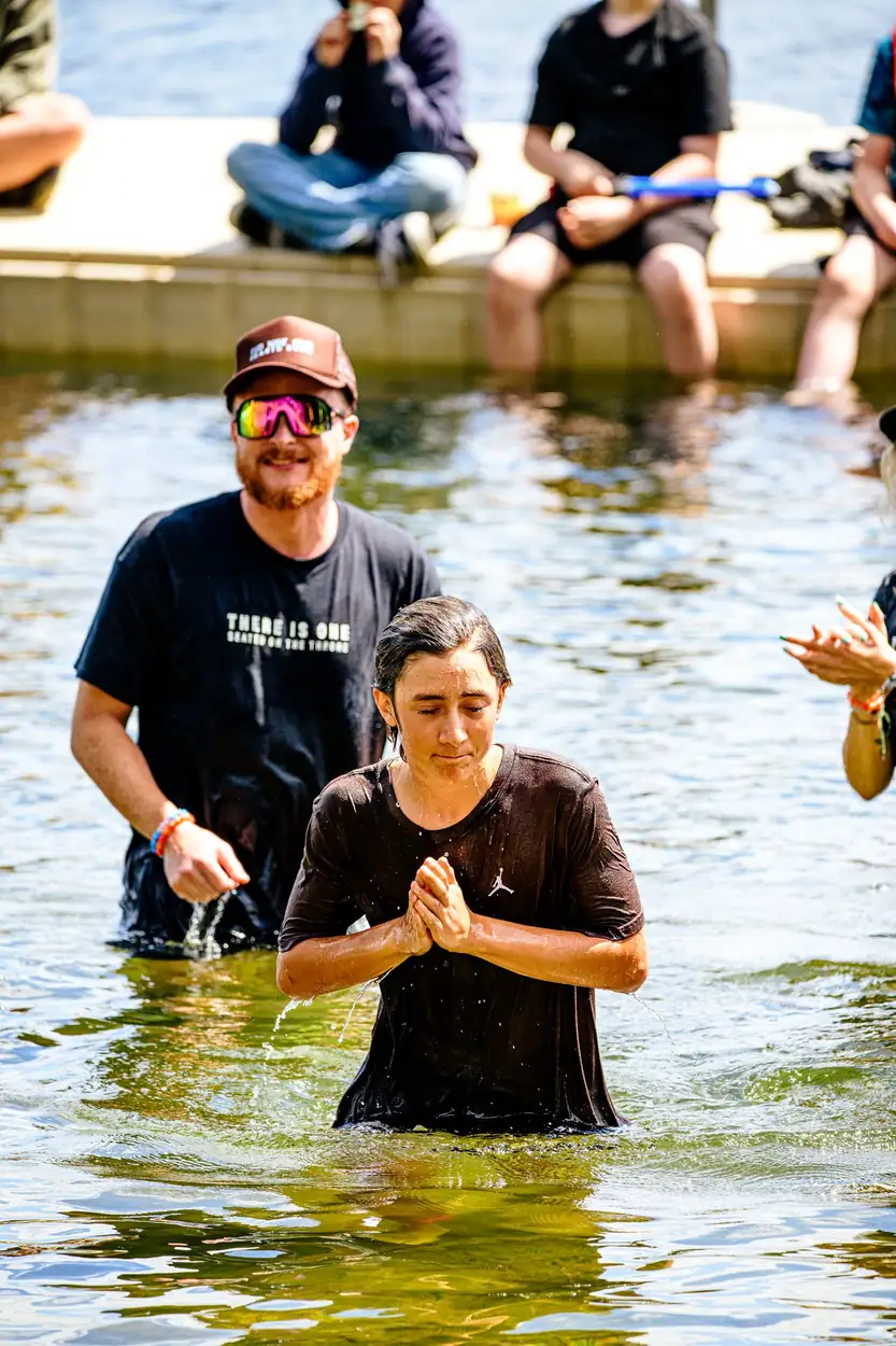 A woman standing waist-deep in the lake, soaked, hands clasped and eyes down, the pastor behind her.
