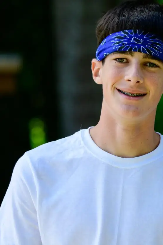 A boy in a plain white tee and blue headband with a quiet half-smile in dappled woods.