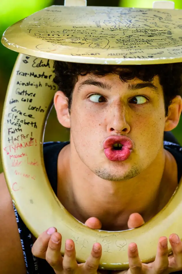 A camper holding a signed wooden toilet seat around his own face like a picture frame, lips pursed, the week's names written all over it.