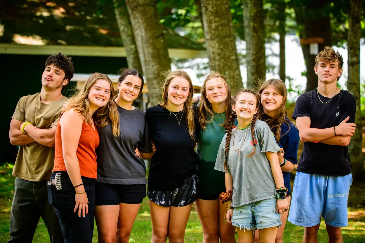 Eight teenagers lined up in the pines with their arms around each other, all smiling at the camera.