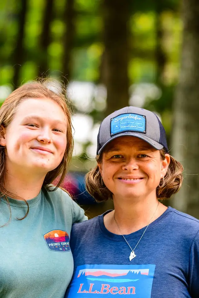 Two women leaders side by side in the pines, both smiling at the camera.