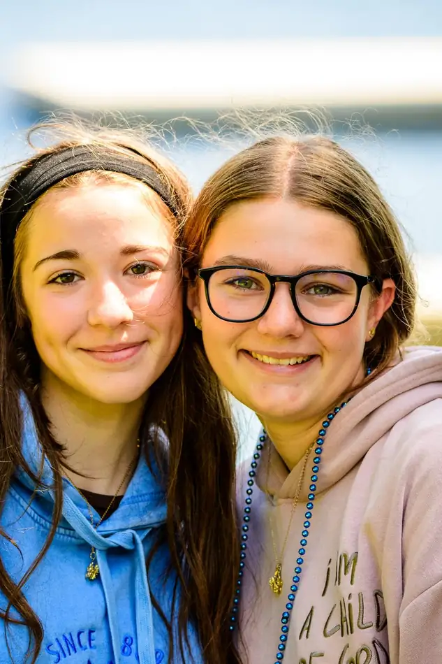 Two girls cheek to cheek, one in a pink hoodie reading I AM A CHILD OF GOD, both in blue bead necklaces.
