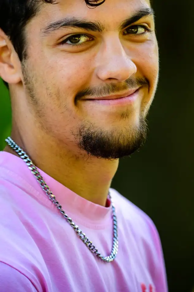 A young man with dark curly hair and a small goatee in a pink tee, smiling.