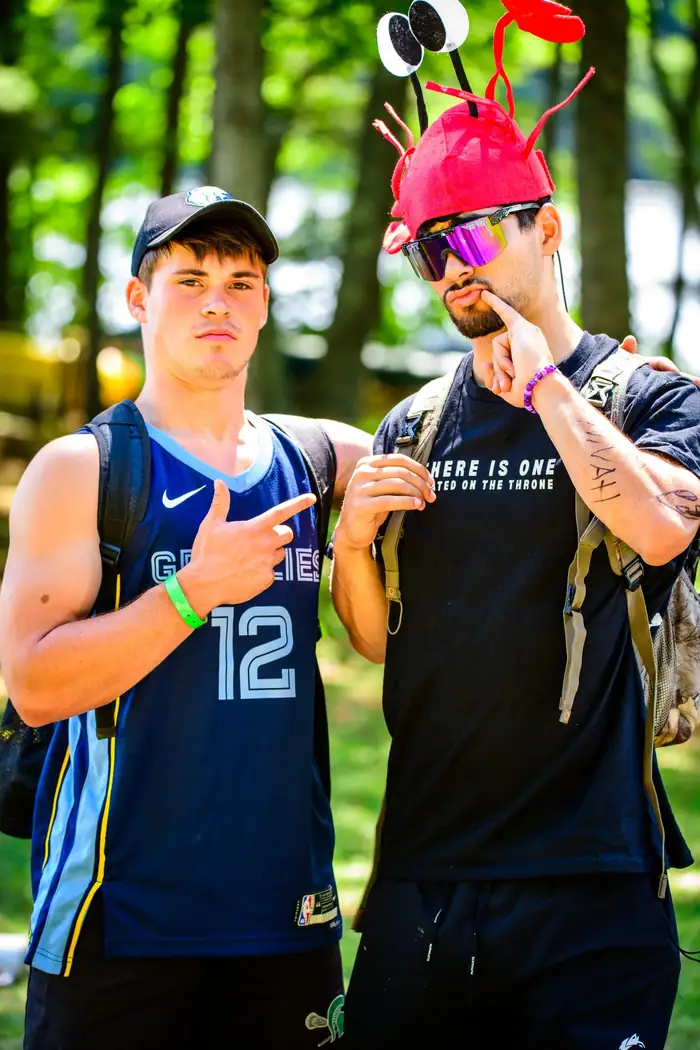 Two campers mugging for the camera, one in a red plastic lobster hat pointing at the writing on his own shirt.